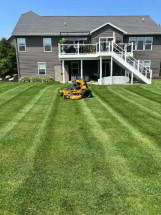 A yellow riding mower cuts striped grass in a backyard behind a gray two-story house with a white deck.